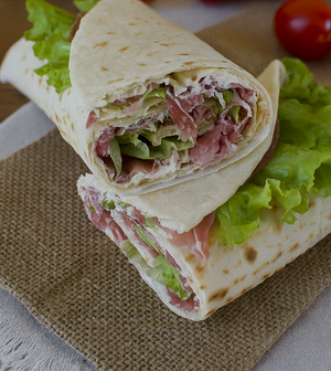 Italian flatbread wrap (piadina) sliced into pinwheels, filled with prosciutto, creamy cheese, and fresh lettuce, arranged on a wooden board with a side of tomatoes and greens